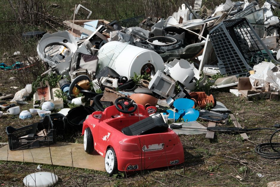 In the foreground of the image, there is a small red plastic toy car with black interior components, situated on a patch of earth and slightly worn with visible scratches and scuffs. Surrounding the toy car are various discarded items, including a black plastic crate, a blue plastic balloon, and tangled black cables. Behind the toy, a large pile of miscellaneous household waste covers a significant area, consisting of broken white appliances, such as washing machine drums and oven parts, with some components showing rust and discoloration. The pile also contains plastic furniture pieces, including panels and frames, as well as cardboard boxes and shredded paper. The arrangement suggests an on-site clearance or an independent waste disposal activity. The background includes overgrown vegetation, with shrubs and small trees framing the scene, indicating an outdoor environment. Natural daylight illuminates the scene, highlighting the textures of the various materials, while the overall setting reflects a typical example of a rubbish removal operation handled by a company like Rubbish Removal Sutton, emphasizing the collection of assorted unwanted household rubbish in a rural or semi-natural area.