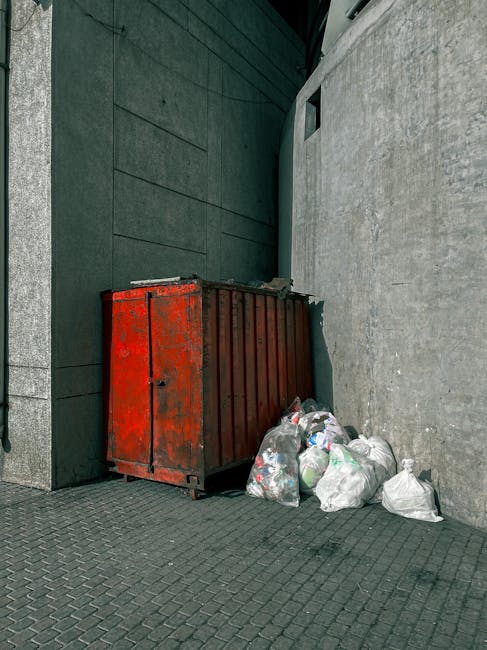 A red metal waste container with visible rust and peeling paint is placed against a dark green building wall and a light gray concrete wall in an outdoor urban setting. Several plastic garbage bags filled with mixed waste are piled beside the container, resting on a paved sidewalk made of dark gray interlocking bricks. The scene is lit with natural daylight, casting soft shadows, and suggests a private or independent waste disposal area for rubbish removal services. The environment appears clean apart from the discarded waste, indicating a site where Rubbish Removal Sutton might handle on-site clearance or private rubbish collection, with the container set to facilitate rubbish disposal near Honeywood Museum or similar locations.