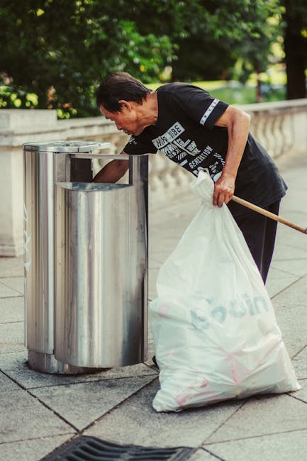 A middle-aged man with short, dark hair and light skin is standing outdoors on a paved walkway lined with a decorative stone balustrade and lush green trees in the background. He is wearing a black T-shirt with white graphic text and dark trousers. The man is holding a long-handled wooden stick in his right hand and is bending slightly forward, reaching into a tall, stainless steel trash bin with a rounded, smooth surface and a rectangular opening at the top, which is integrated into a cluster of three similar bins. In his left hand, he holds a large white plastic garbage bag, which appears partially filled with waste. The setting suggests an area for public waste disposal, and the scene is evenly lit with natural daylight, emphasizing the cleanliness and practicality of the waste management activity and subtly aligning with the services offered by Rubbish Removal Sutton for private or community rubbish clearance.