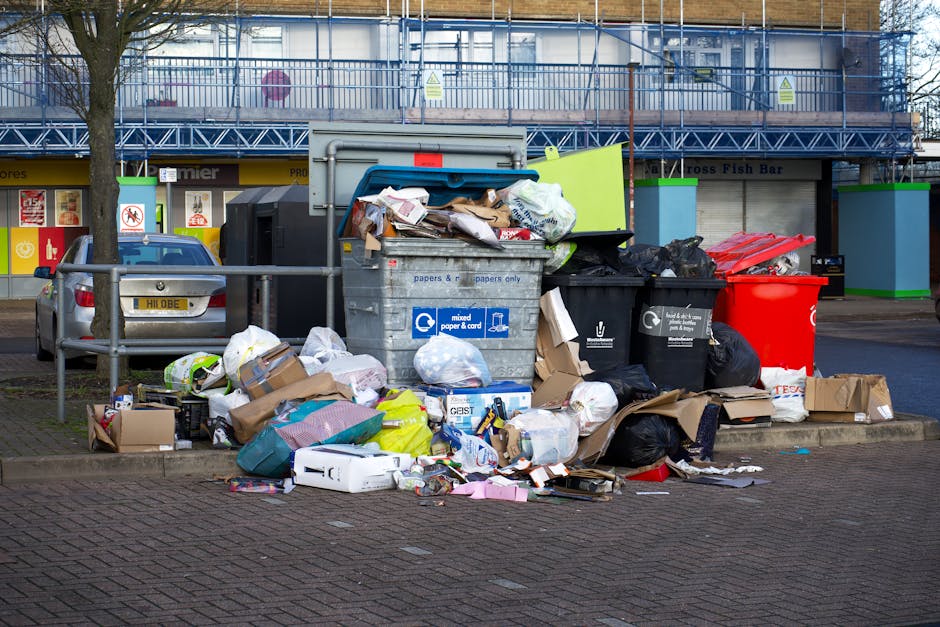 A large collection of mixed waste, including black and black-and-white garbage bags, flattened cardboard boxes, and loose paper and packaging, is piled on a paved area in front of a multi-story building under construction, which is covered in blue scaffolding. The waste also includes plastic containers, a red wheelie bin, a black recycling bin, and a green bin, all situated on the sidewalk next to a parked silver car. The background features retail units with signage, and a tree partially obstructs the view on the left. The scene appears to depict an overflow of rubbish awaiting collection, illustrating an example of private waste handling or rubbish removal service, such as that provided by Rubbish Removal Sutton, for clients needing alternative solutions outside standard local authority collection. The overall lighting is natural, with a slightly overcast sky, emphasizing the cluttered and unmanaged appearance of the waste.