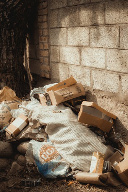 A large pile of black plastic rubbish bags stacked against a modern building with a tiled exterior wall. Some of the bags are partially torn, revealing white waste and plastic bottles inside. Several transparent plastic bottles are visible, some with caps, mixed among the bags. The bags are unevenly piled, with a few leaning or partially open, exposing rubbish beneath. In the background, a section of the building's facade shows a graffiti tag in red spray paint that reads 'XEND.' The scene appears to be outdoors, possibly on a paved area or street, under diffuse natural lighting that highlights the textures and shiny surfaces of the bags. This image relates to private waste collection or alternative rubbish removal services such as those offered by Rubbish Removal Sutton, illustrating unmanaged or excess waste awaiting collection near an urban setting.