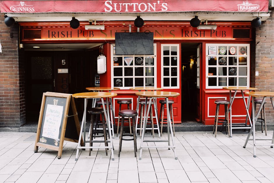 The exterior of a traditional Irish pub features a red-painted façade with large white-framed windows and a wooden door behind the outdoor seating area. In front of the pub, there are three round wooden high tables with metal legs, accompanied by multiple black bar stools with round seats, arranged on a paved sidewalk. A small A-frame chalkboard sign stands on the pavement, displaying handwritten menu items or notices. Above the entrance, a black flat-screen television is mounted, and a white electric box and other small fixtures are visible on the wall beside the doorway. The pub's signage includes the words