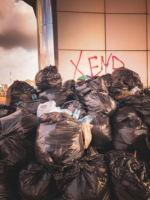A large pile of black bin bags filled with rubbish is situated against a tiled wall outdoors, with some bags appearing partially torn or bulging, revealing contents such as plastic bottles and waste materials. The bags are arranged haphazardly, with a few leaning or stacked irregularly. Behind the pile, the wall features a red spray-painted word 'XEND' on the light-colored tiles, and part of a reflective metallic surface, possibly a cylindrical column, is visible to the left. The scene is set in a public or urban environment, with an overcast sky in the background that creates diffuse lighting, emphasizing the textures of the plastic bags and the environmental setting. This scene exemplifies private waste accumulation that may be associated with alternative rubbish removal or on-site clearance services, often handled by independent waste collection providers like Rubbish Removal Sutton, reflecting how non-local authority disposal methods can differ from municipal waste management.